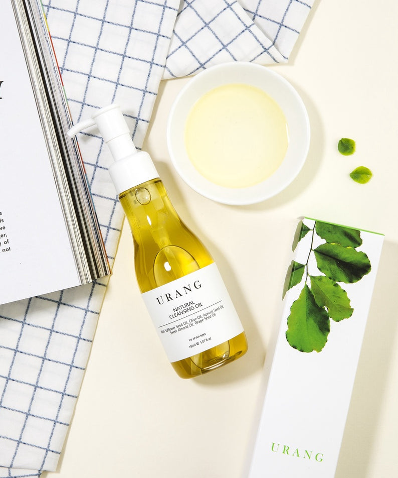 Flacon de URANG Natural Cleansing Oil 150ml sur fond doux avec boîte et ingrédients naturels.