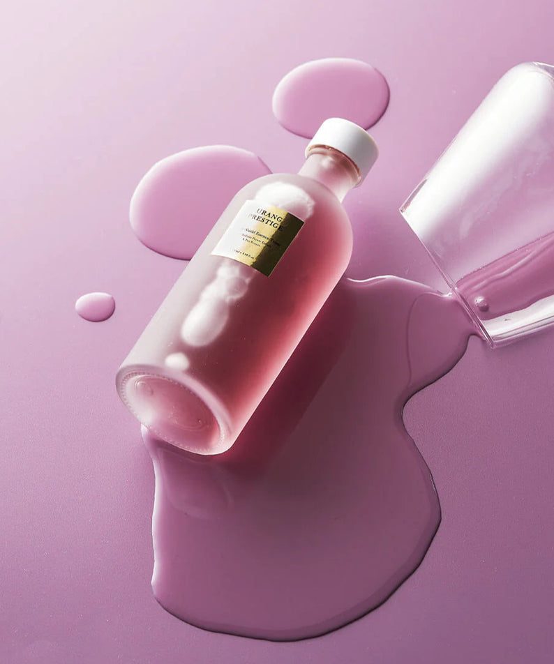 Flacon de l'URANG Prestige Purple Shield Essence Toner sur fond lavande, entouré de gouttes, essence délicate pour la peau.