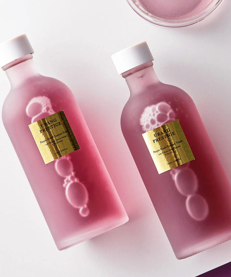 Deux bouteilles de URANG Prestige Purple Shield Essence Toner sur fond rose.