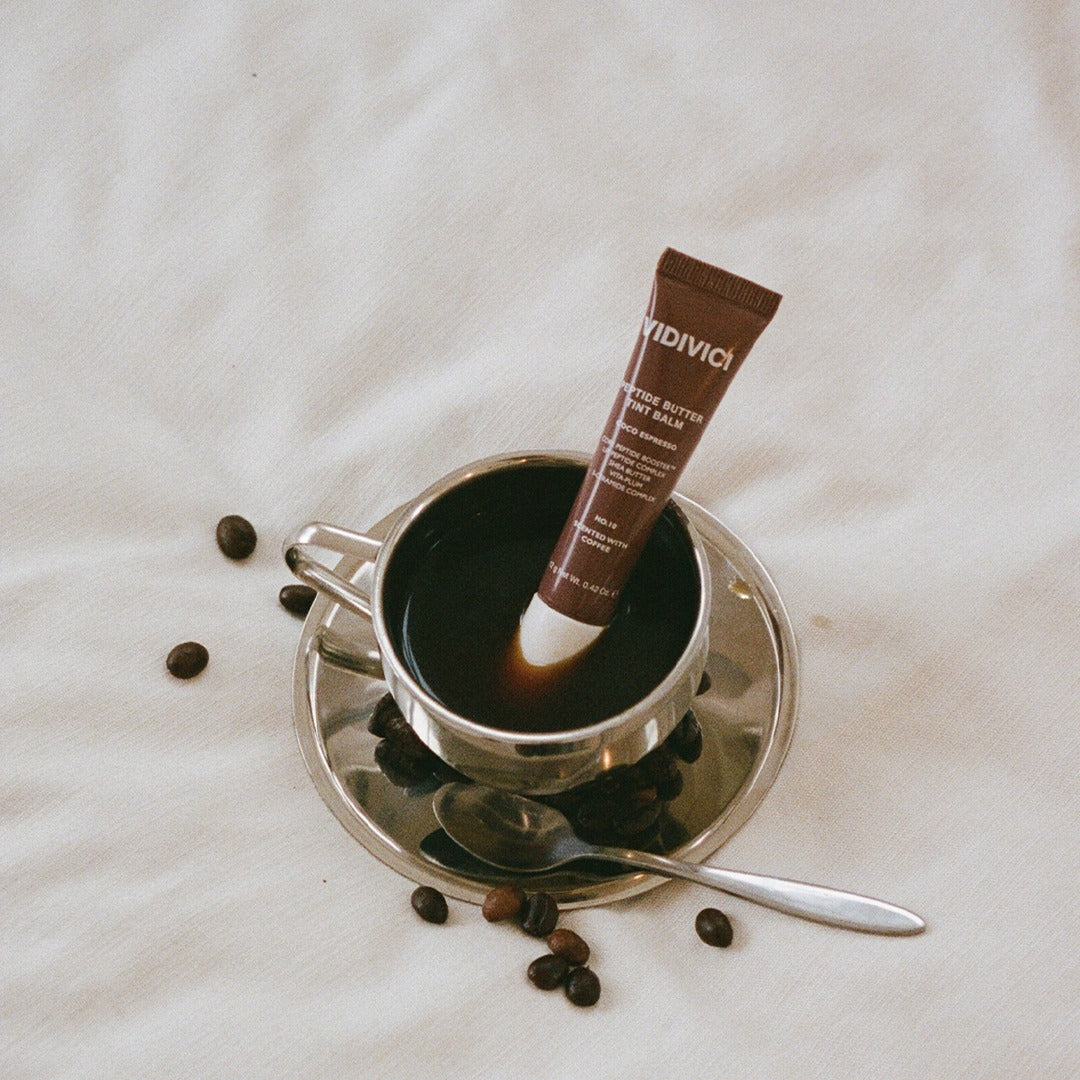 VIDIVICI Peptide Butter Tint Balm de la marque VIDIVICI, tube brun sur tissu clair, mise en scène près d’une tasse et grains.