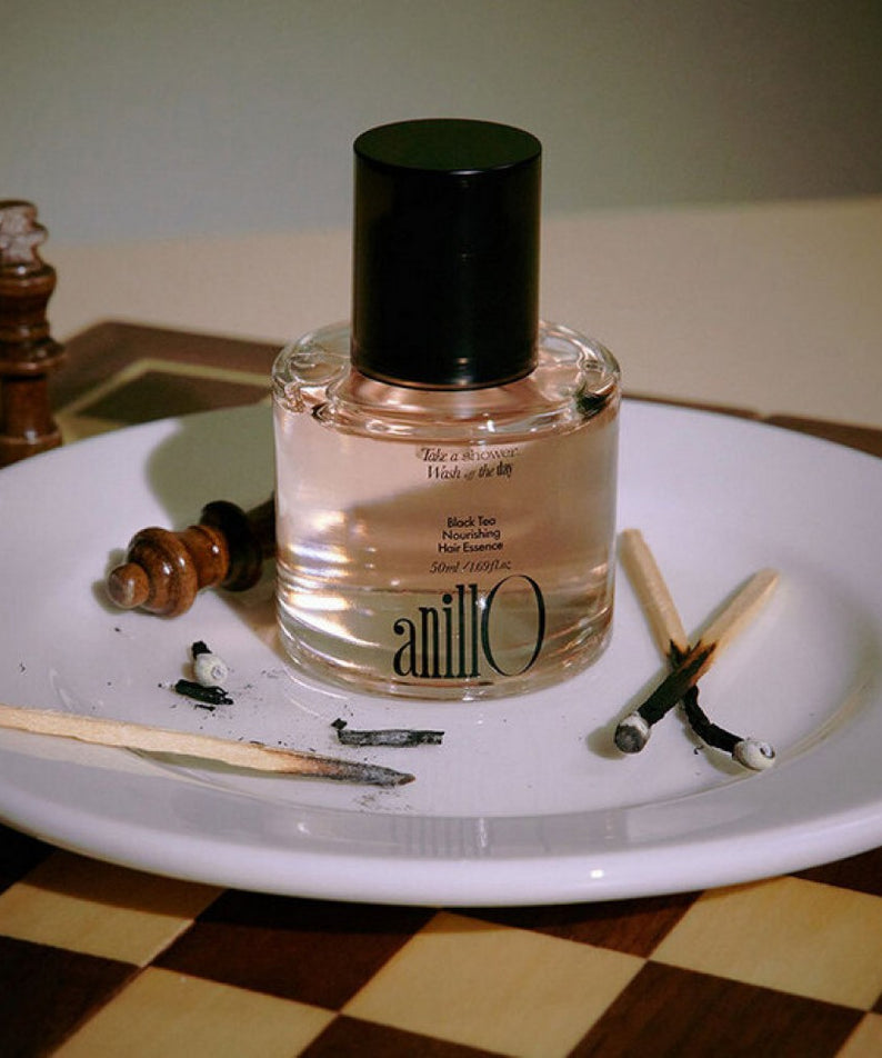 ANILLO Black Tea Nourishing Hair Essence 50ml : flacon noir et ambre sur une assiette, décor salon cosmétique, ambiance naturelle.
