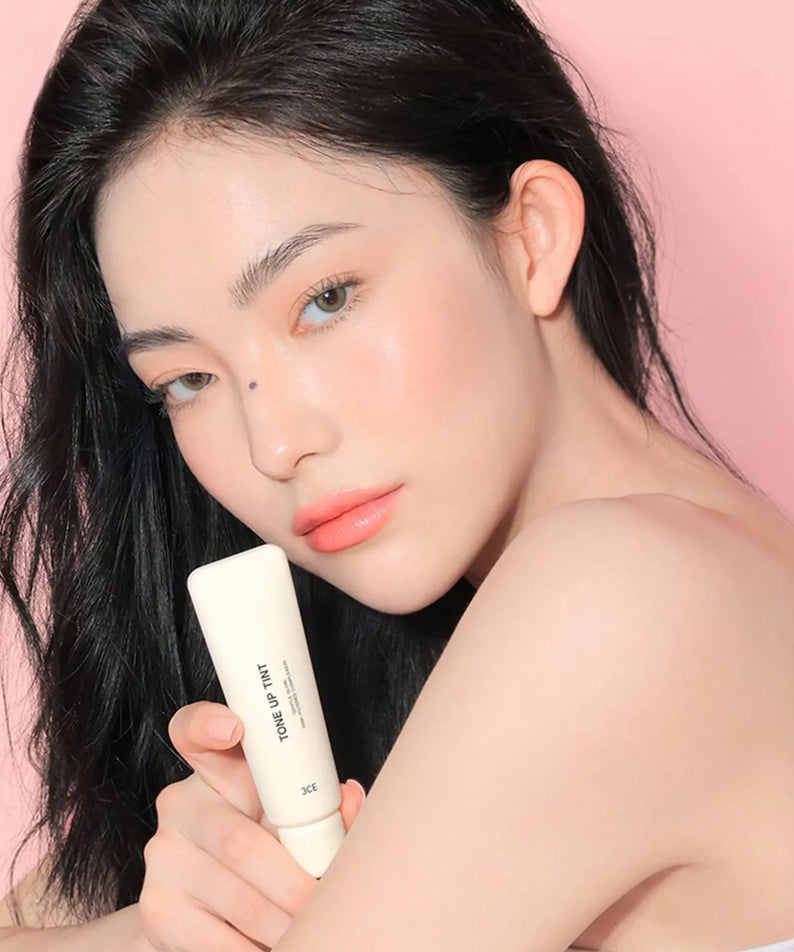 Décris l'image en français : 3CE Tone Up Tint 40ml STYLE NANDA, tube crème sur peau avec jeune femme maquillée fond rose.