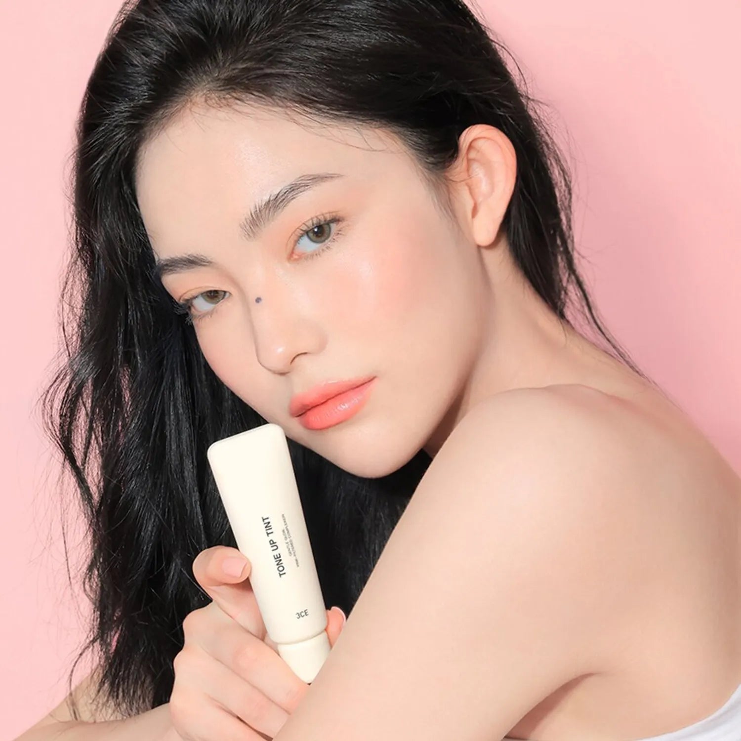 Décris l'image en français : 3CE Tone Up Tint 40ml STYLE NANDA, tube crème sur peau avec jeune femme maquillée fond rose.