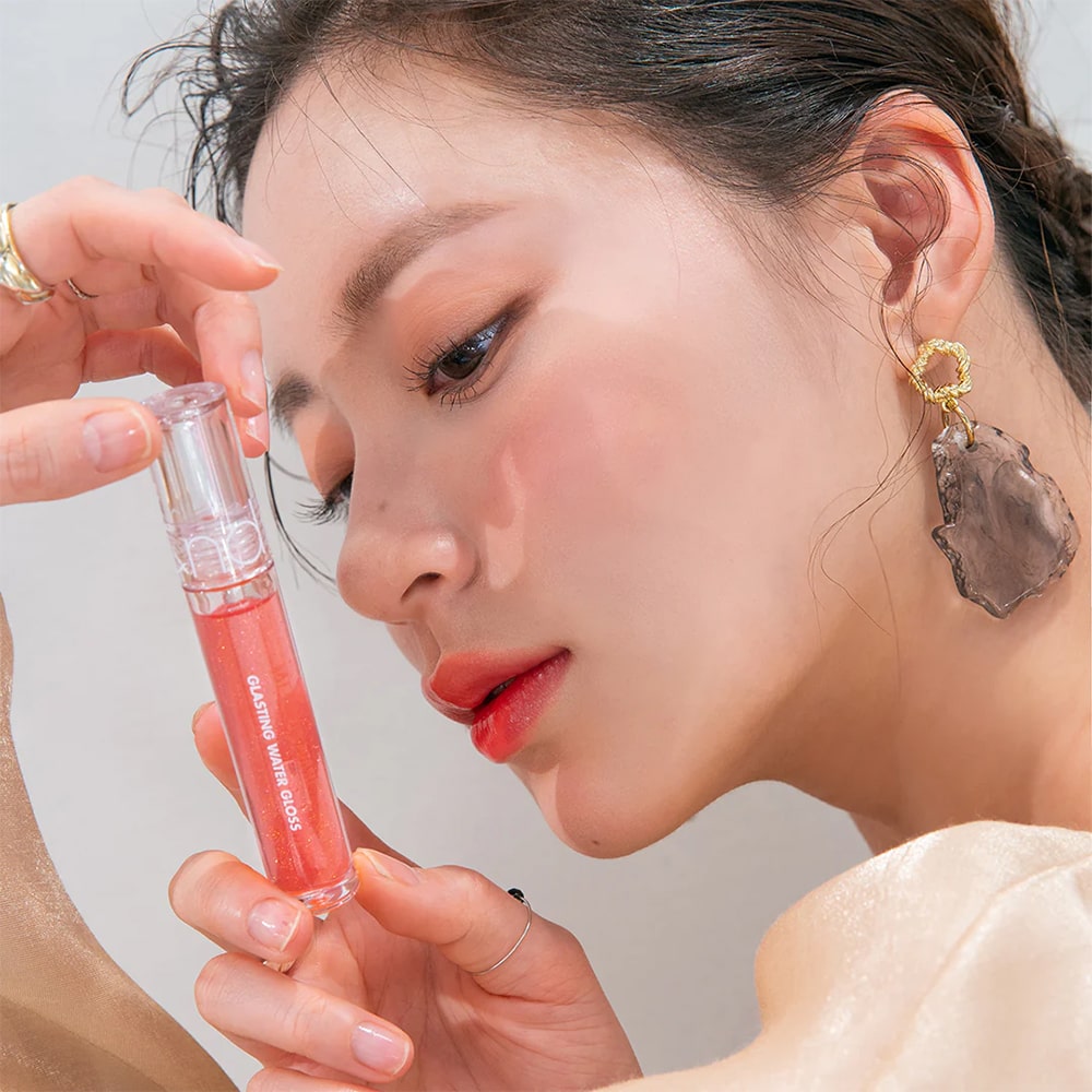 ROMAND Glasting Water Gloss, flacon rosé appliqué sur le maquillage, gros plan visage en studio, main à côté.